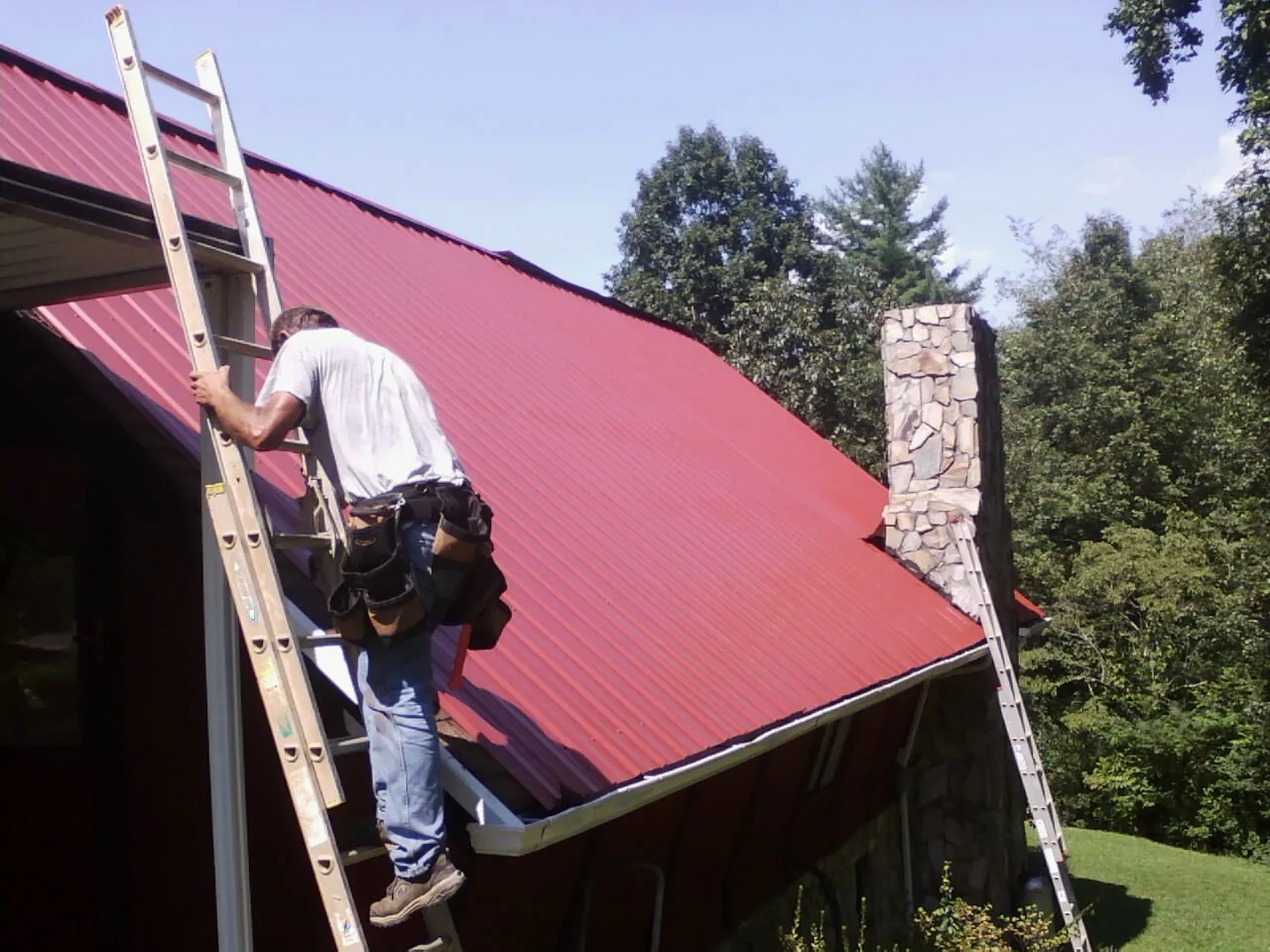 Roofer on a ladder beside a red metal roof and stone chimney — Soffit & Fascia Repair by Roofing Brothers in Latah